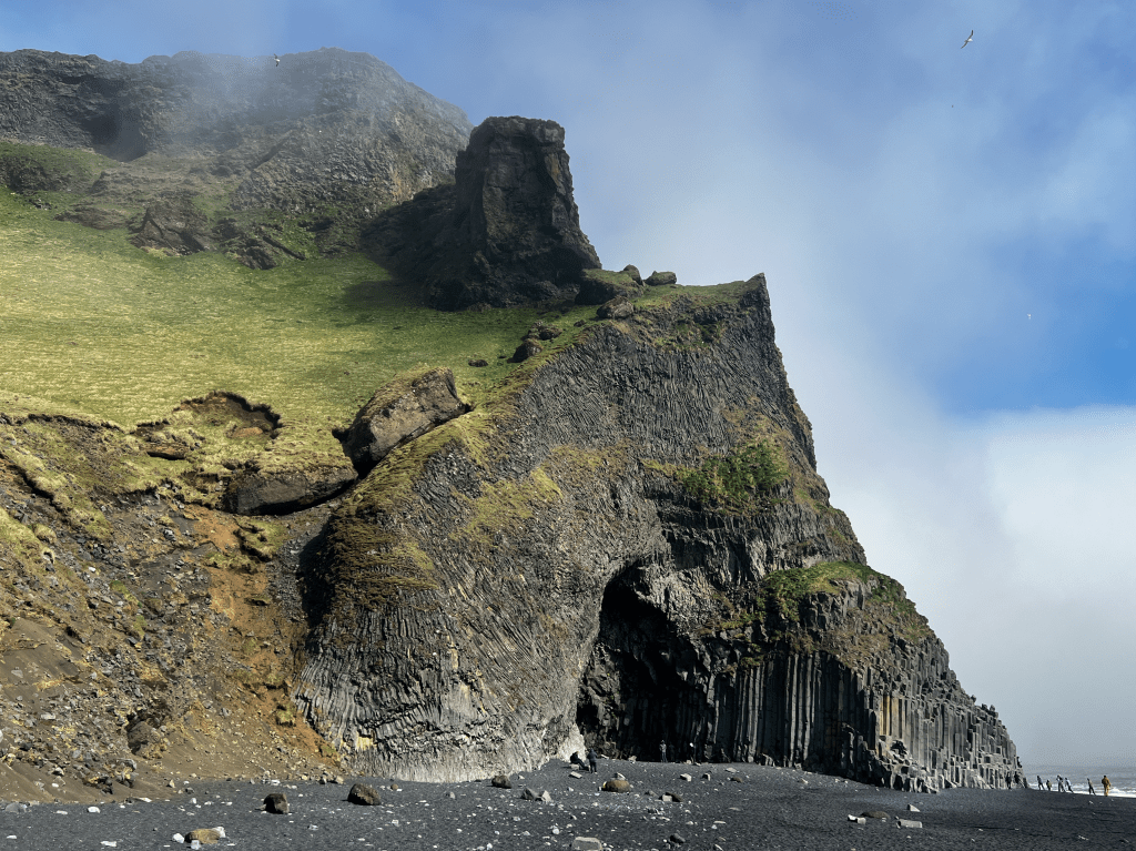 A rocky cliff covered with grass rises above a black sand beach, partially obscured by fog.