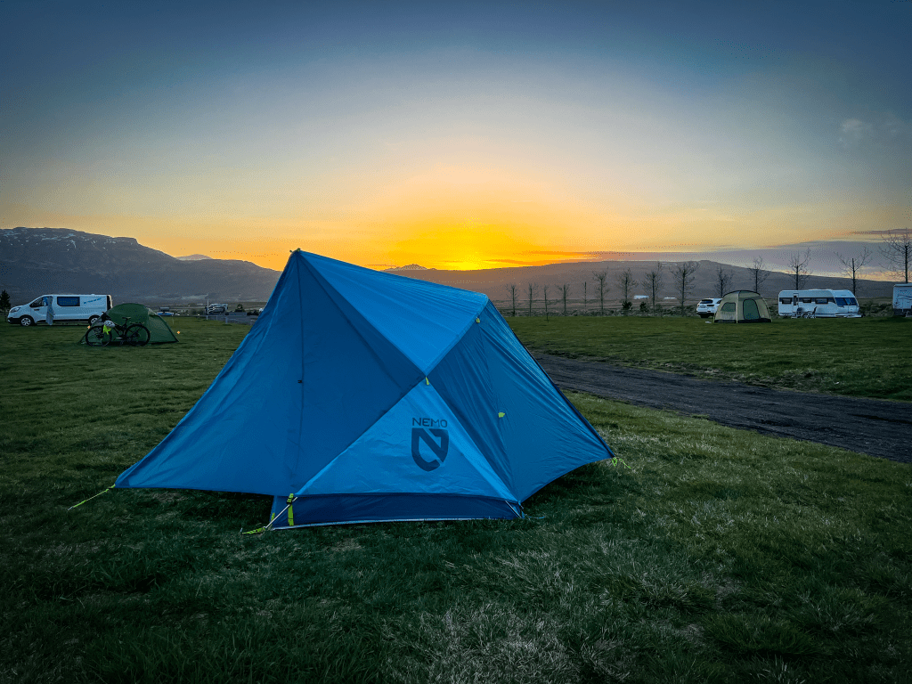 A blue camping tent set against a sunset backdrop at a campsite in Iceland, with other tents and vehicles visible in the distance.