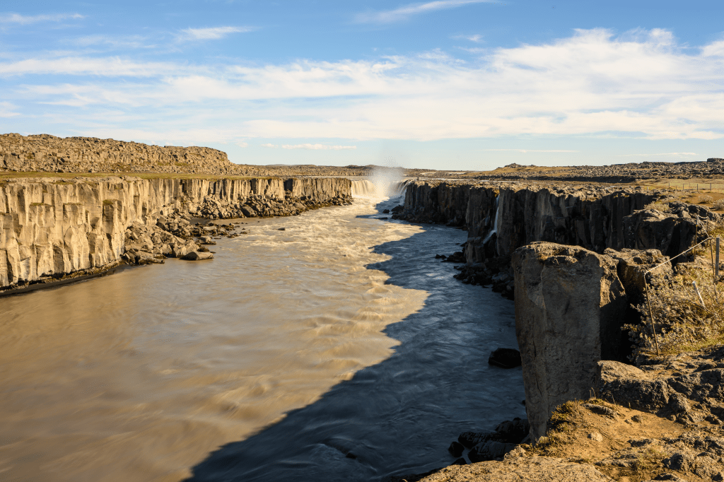 A scenic view of a river flowing between rocky cliffs, with a waterfall cascading in the distance under a clear blue sky.