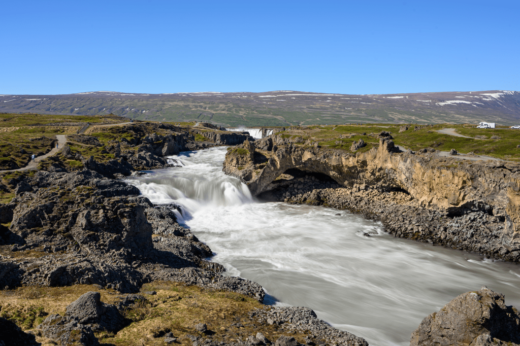 A panoramic view of a rushing river flowing over rocks, with a waterfall visible in the distance under a clear blue sky.