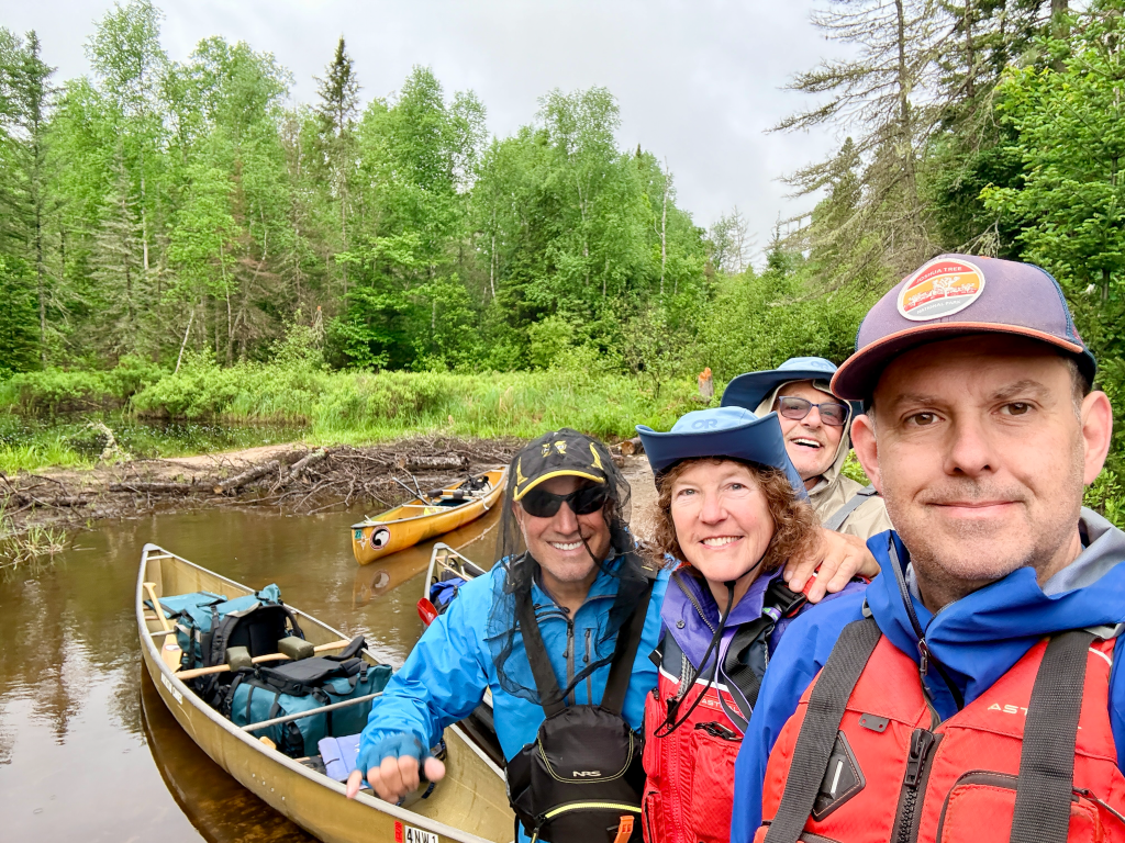 Group of four people smiling together by the water, with canoes in the background and lush green trees surrounding them.
