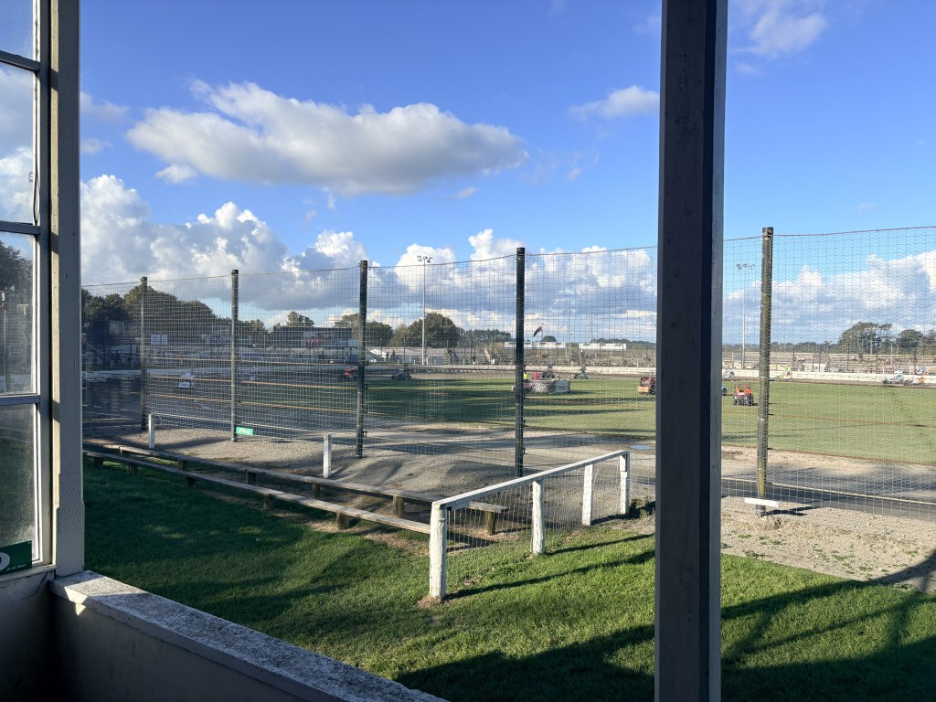 A view of a racetrack from a seating area, featuring a fenced perimeter, grassy areas, and vehicles in the distance under a cloudy sky.
